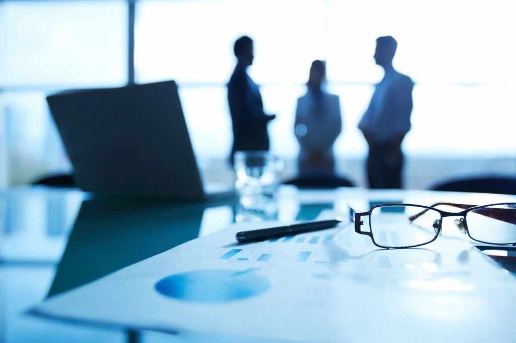 A blurred office scene with documents, glasses, and three people in silhouette discussing in the background.